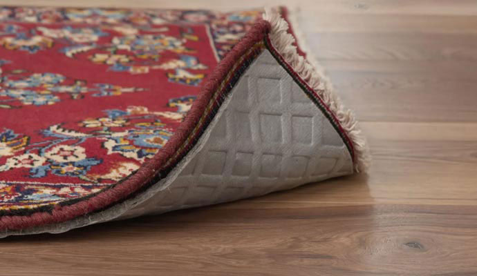 Corner of a red oriental rug lifted to show a protective non-slip rug pad on a hardwood floor